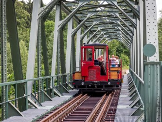 Das Bild zeigt eine Lok der Chemnitztalbahn auf Gleisen, welche gerade eine Brücke überquert. In einem angehängten Waggon sitzen Menschen.