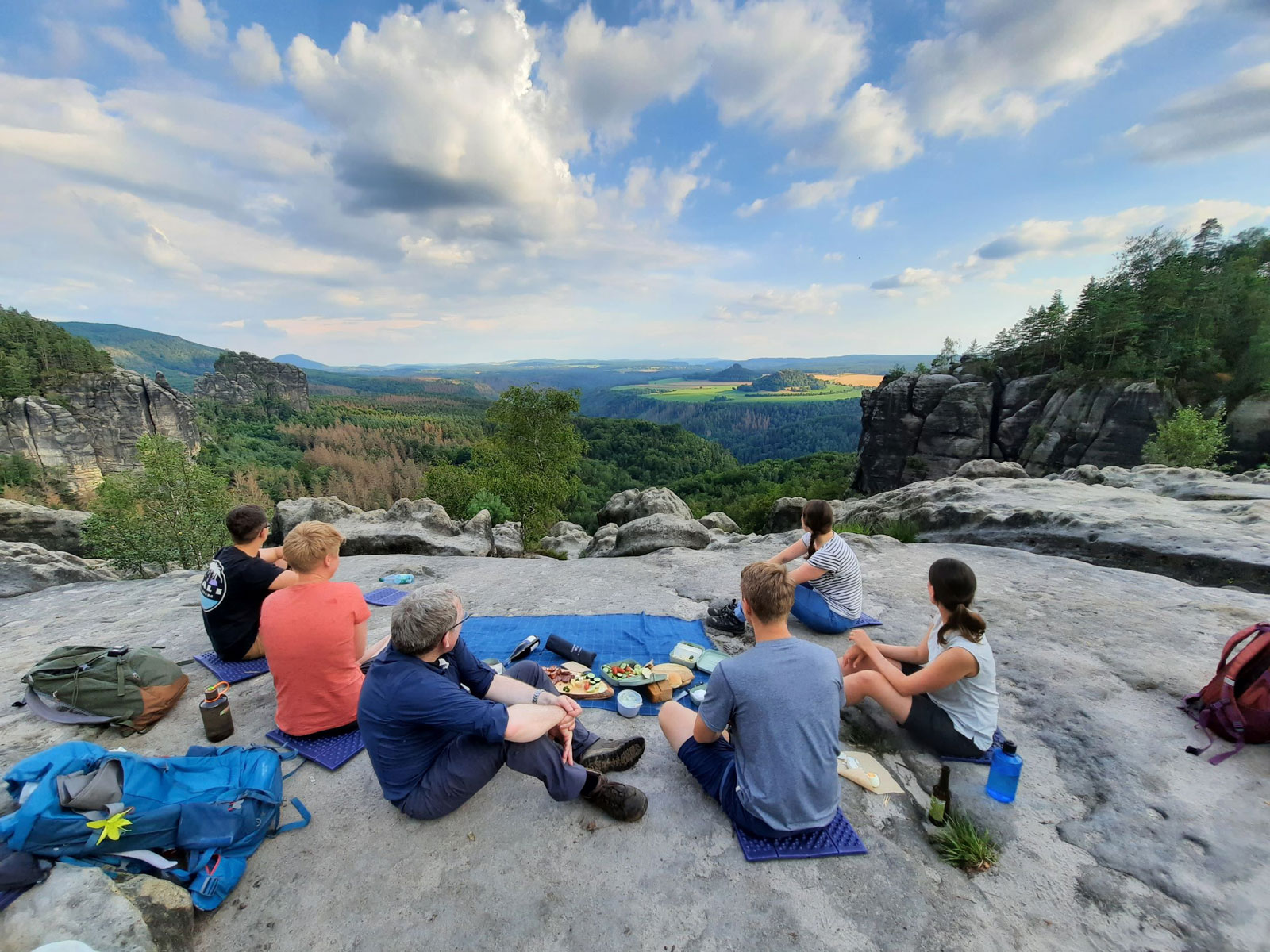 Sechs Personen machen ein Picknick hoch oben auf dem Berg und gucken in die Ferne.