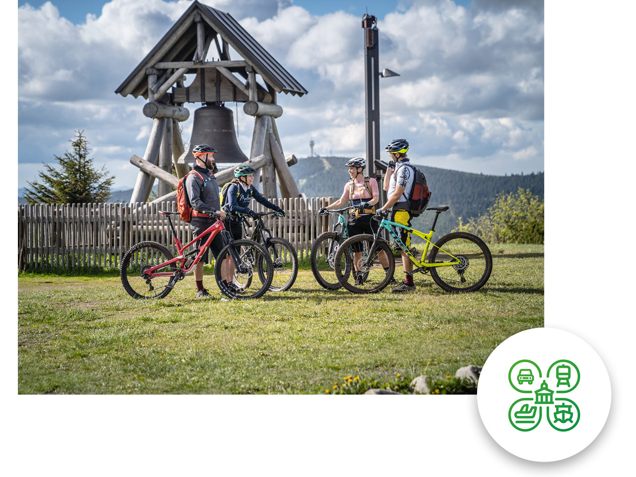 Vier Fahrradfahrer:innen stehen mit ihren Mountainbikes am Gipfel Fichtelberg vor einer Glocke (Denkmal).