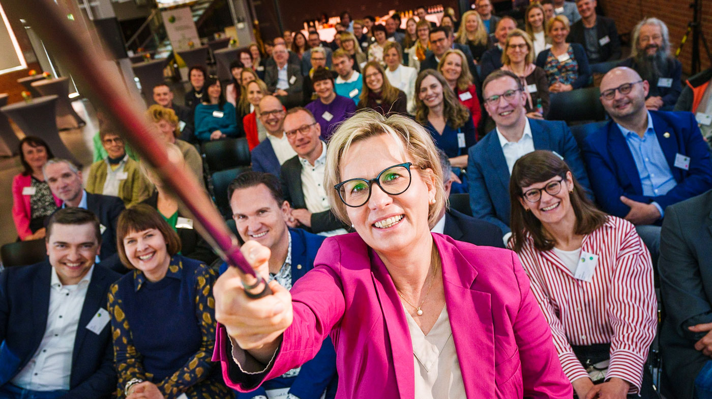 Ministerin Barbarba Klepsch hält einen Stelfiestick in der Hand.Im Hintergrund sitzen ganz viele Menschen und schauen freundlich in die Kamera.